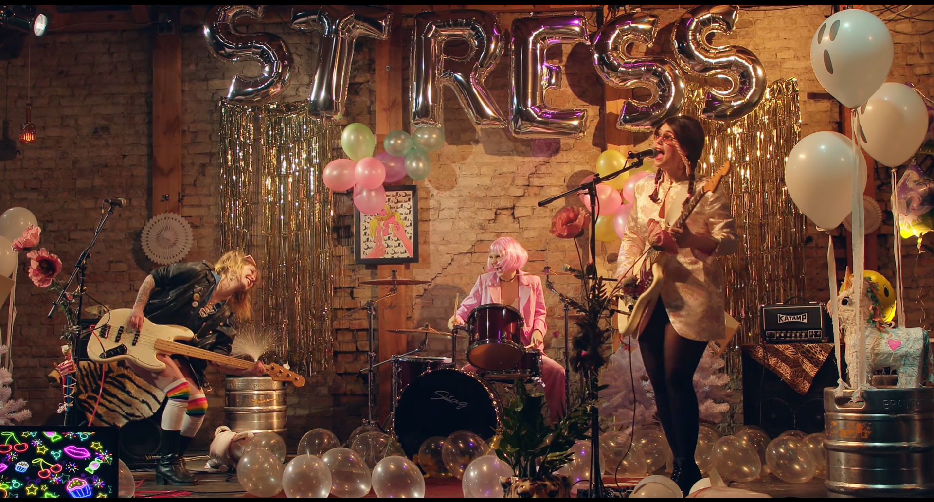 Kat, Mary e Karo no videoclipe "Bitter Lollipop". Cenário de festa, com balões ao fundo escrito "Stress" enquanto o grupo toca.
